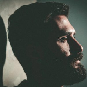 The profile view of a bearded man looking ahead with a dark background as he considers what type of addiction treatment center is best for him.