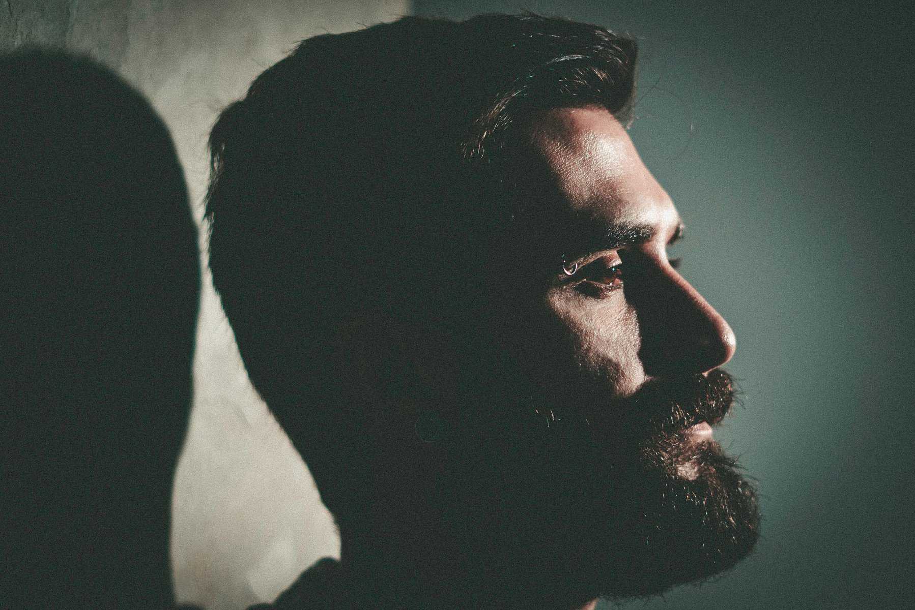 The profile view of a bearded man looking ahead with a dark background as he considers what type of addiction treatment center is best for him.